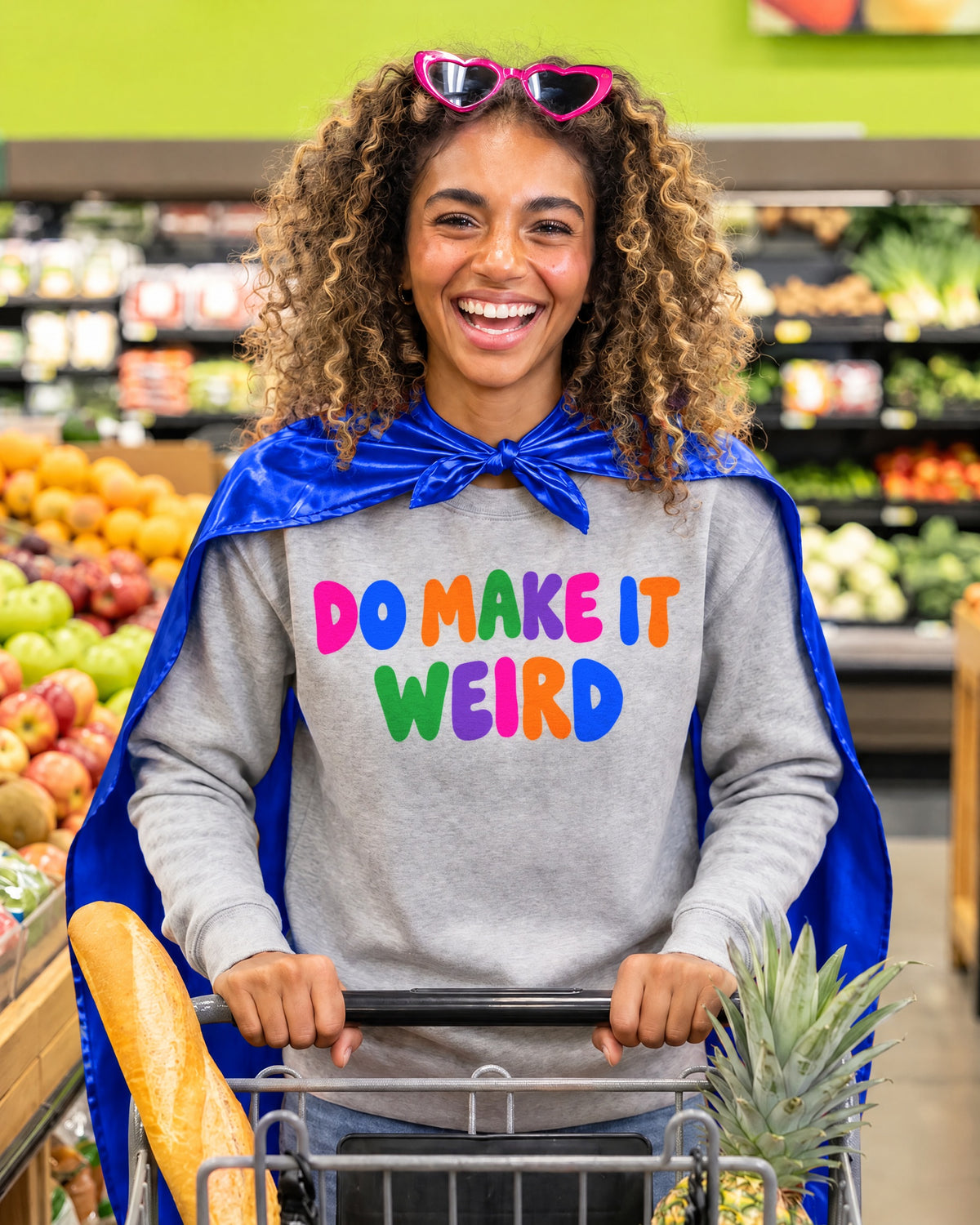 AI-generated image: a woman in a blue superhero cape pushing a grocery cart, wearing the ash Do Make It Weird sweatshirt